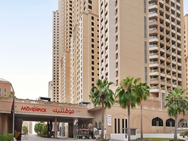 Exterior view of tall beige residential towers above a Mövenpick entrance archway, with palm trees and a driveway in the foreground.