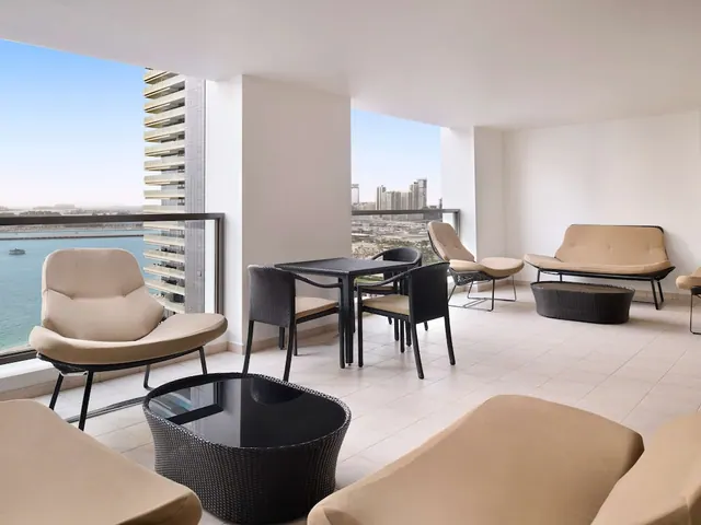A covered balcony with cream lounge chairs, a black wicker table and chairs, and views of the sea and city towers.