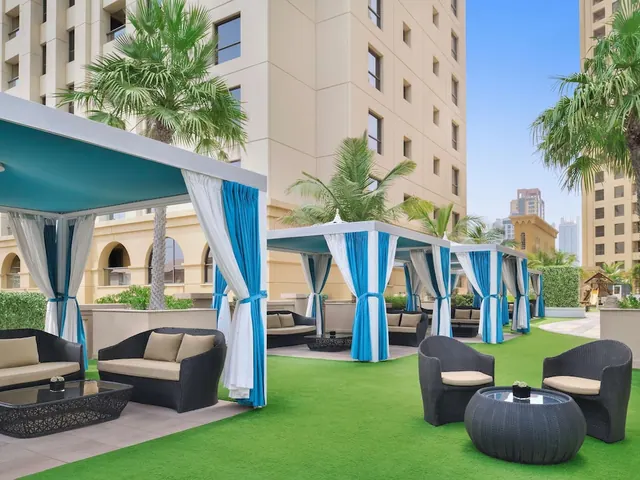 Outdoor lounge area with blue and white curtained cabanas, wicker seating and artificial turf, with palm trees and a high-rise building behind.
