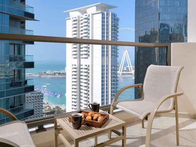A balcony with two beige chairs and a small table holding a tray of croissants and two coffee mugs, overlooking the sea, high-rise towers and the Ain Dubai ferris wheel.
