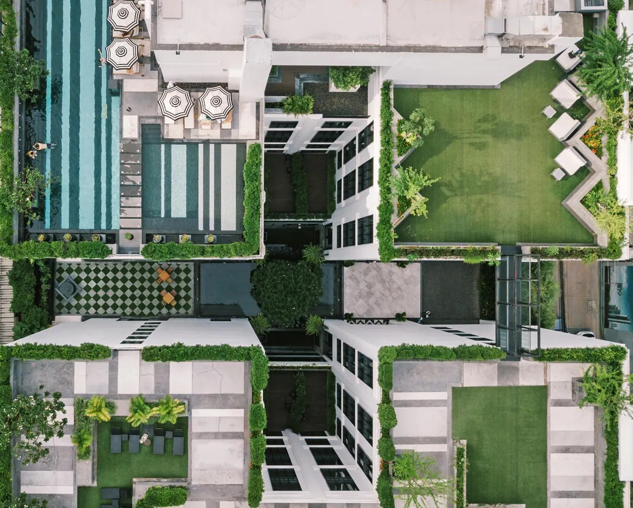 Aerial view of a hotel complex with two swimming pools, striped umbrellas, lawns, and loungers arranged around internal courtyards.