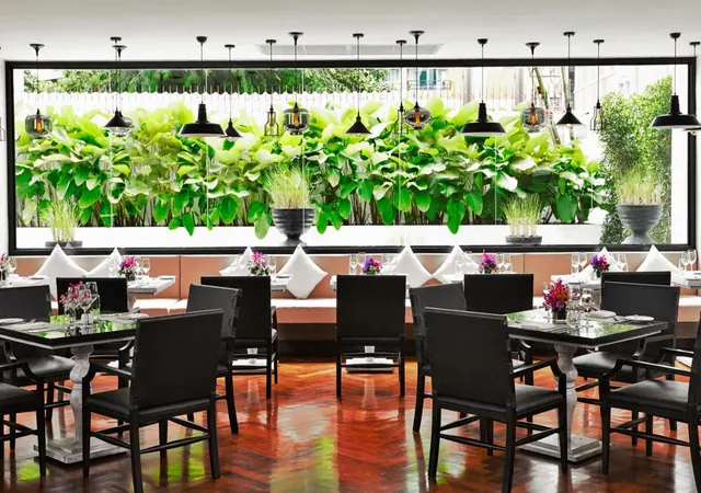 A restaurant dining room with black chairs and glass-topped tables under pendant lights, with a large window looking out onto green plants and trees.