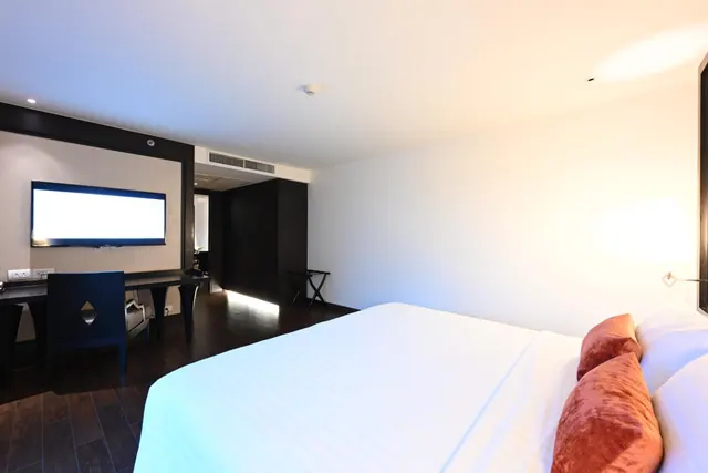 A modern hotel bedroom with a made white bed with rust-coloured cushions, a wall-mounted television and a work desk.