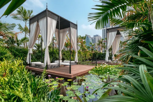 A black metal canopy daybed with white curtains sits on a timber deck beside a pond of water lilies, with tall buildings visible behind the tropical garden.