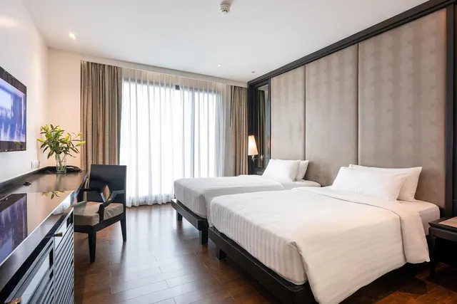 A hotel bedroom with two single beds with white bedding, a wall-mounted television, a dark desk and chair, and sheer curtains over a bright window.