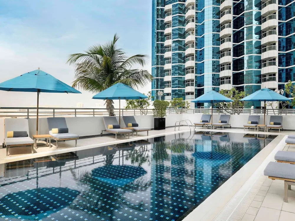 A rooftop swimming pool with blue umbrellas and sun loungers sits beside a tall glass tower with balconies and a palm tree.