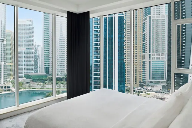 A bedroom with a made white bed facing floor-to-ceiling windows that look out over high-rise towers and a body of water with palm trees below.