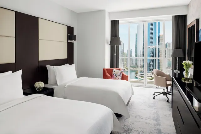 A bedroom with two twin beds, a red armchair, a desk and chair, a wall-mounted television, and floor-to-ceiling windows overlooking high-rise towers.