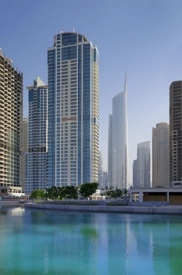 Exterior of a tall glass hotel tower with the Movenpick name on its facade, seen across a canal with neighbouring skyscrapers beside it.