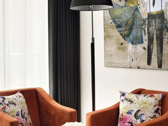 A living area with two rust-orange armchairs with floral cushions, a black floor lamp, dark curtains and a large framed painting.