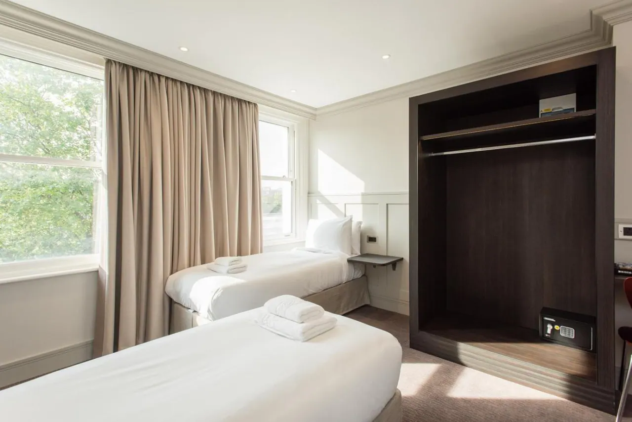A bedroom with two single beds with white linen and folded towels, a window with trees outside, and an open wardrobe with a built-in safe.