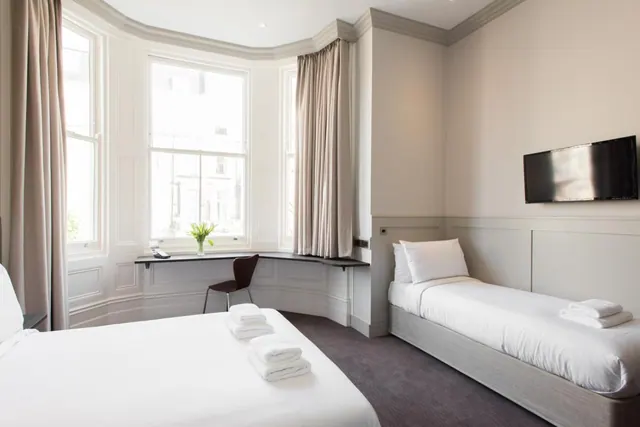 A hotel bedroom with a large bed in the foreground, a single bed on the right, a wall-mounted television, and a desk with a chair beneath a bay window.
