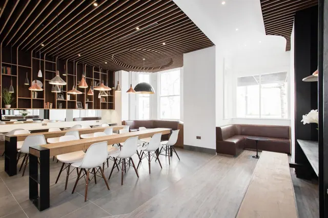 A dining area with long wooden tables, white chairs, leather banquette seating and pendant lights beneath a slatted timber ceiling.
