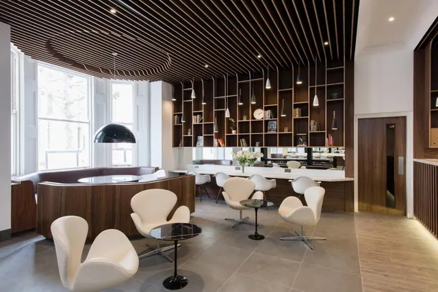 A lobby lounge with white swan chairs, round black tables, a curved wooden banquette by the window, and a slatted wood ceiling above a shelved wall and counter.