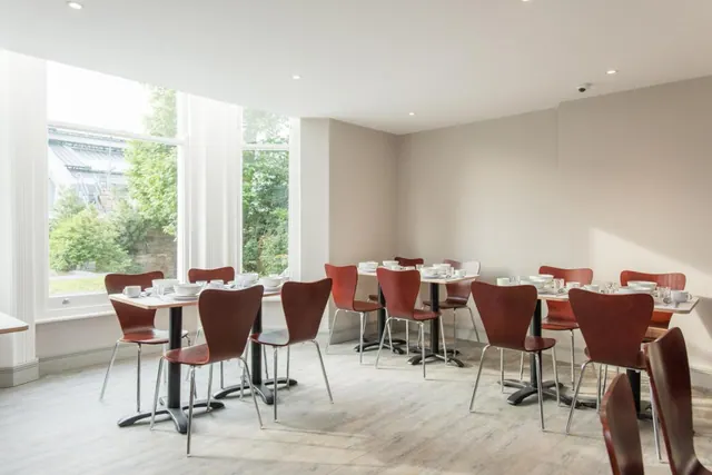 A dining room with brown wooden chairs and set tables beside tall windows that look onto trees and a glass roof outside.