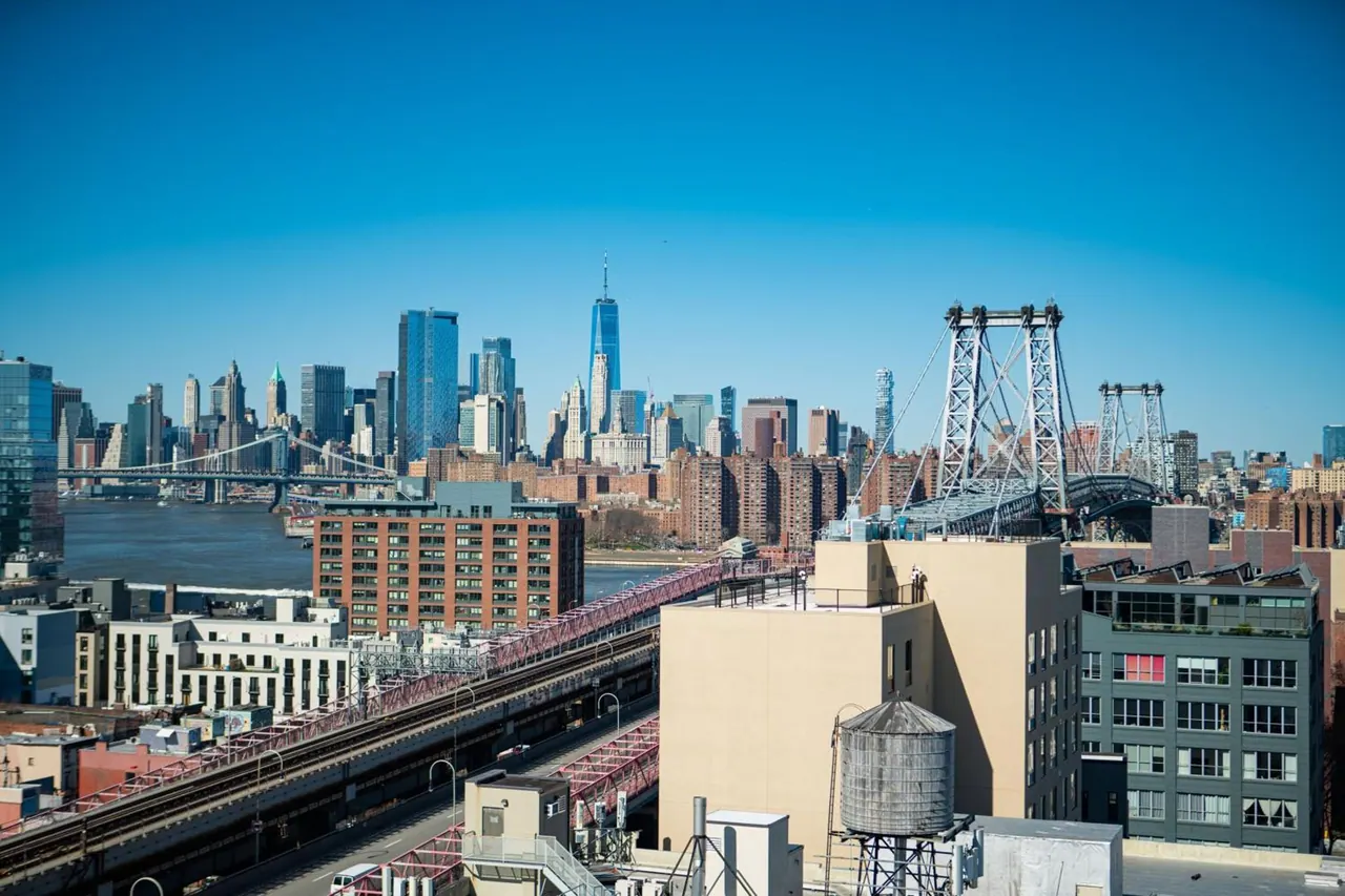 A view over rooftops toward the Lower Manhattan skyline, with One World Trade Center and the Williamsburg Bridge visible under a clear blue sky.