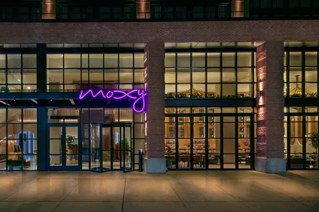The exterior of a brick and glass building at night, with a purple neon Moxy sign above the entrance and a revolving door at street level.