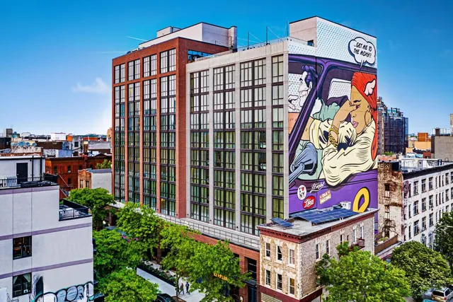 The exterior of a tall hotel building with a large pop-art mural of a comic-style couple and a speech bubble reading 'Get me to the Moxy!!', beside older brick and stone buildings and street trees.