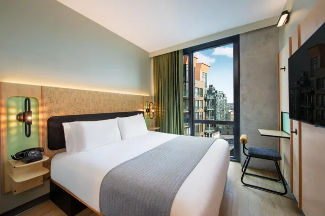 A king bed sits beside a tall window looking out over nearby city buildings, with a wall-mounted television and a small desk with a chair.