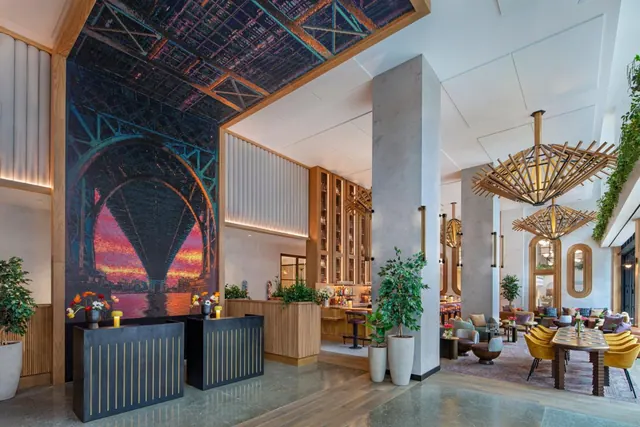A hotel lobby with two dark reception desks in front of a large bridge mural, with a seating lounge and woven pendant lights visible to the right.