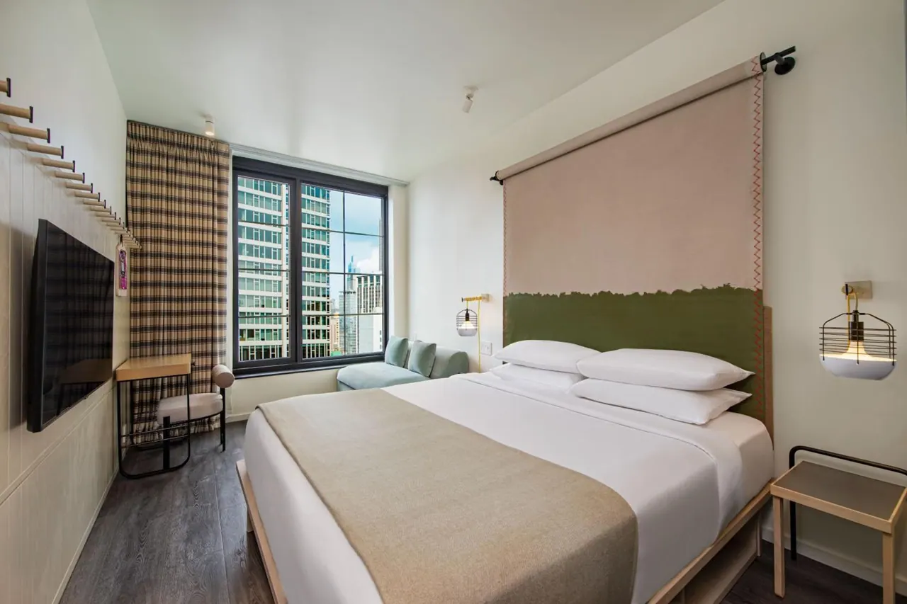 A bedroom with a made king bed, a wall-mounted television, a small desk and chair, and a seating area by a window overlooking tall buildings.