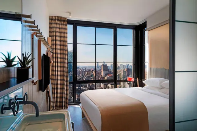 A bedroom with a king bed beside floor-to-ceiling windows that look out over a city skyline.