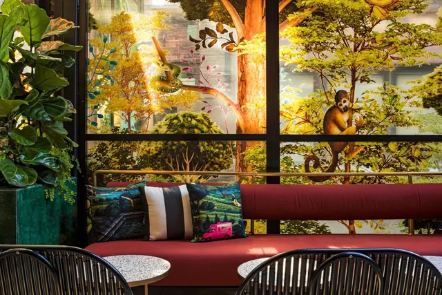 A red banquette with patterned cushions and round terrazzo tables sits in front of large windows decorated with a painted tree mural.