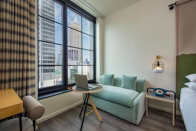 A hotel room with a mint green sofa, a small round table holding a laptop, and a large window view of the Empire State Building.