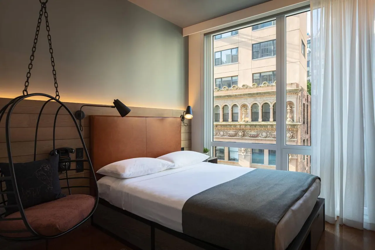 A bedroom with a double bed beside large windows looking onto an ornate stone building across the street, with a hanging chair on the left.