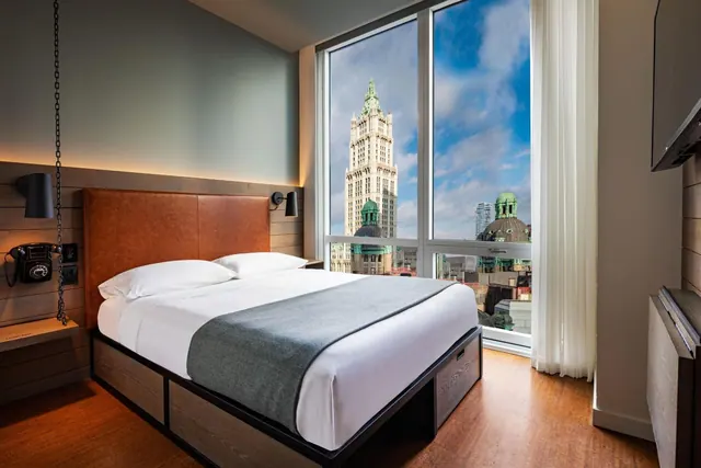 A bedroom with a made bed beside floor-to-ceiling windows showing the Woolworth Building tower and domed rooftops under a blue sky.