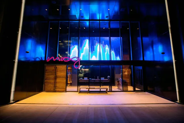 Night view of a hotel entrance with blue-lit glass walls, a pink neon sign reading moxy, wooden doors, and a bench on the paved forecourt.