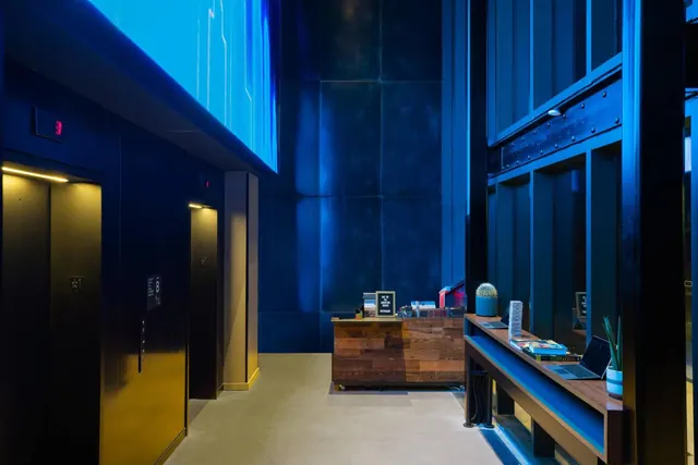 A hotel lobby with two elevator doors on the left, a wooden reception desk in the centre and a wooden console holding a laptop and a cactus on the right, under blue lighting.