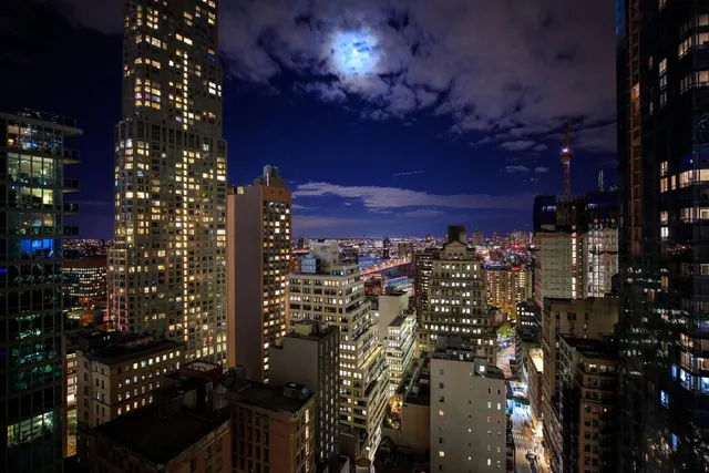 Night view over dense high-rise buildings with many lit windows, a moon behind clouds, and a lit bridge in the distance.