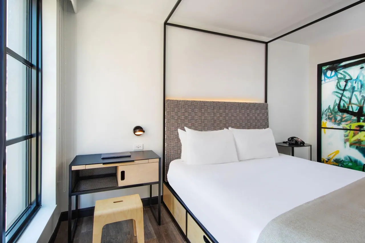 A bedroom with a canopy-style king bed with an upholstered headboard, a black desk with a laptop by the window, a wooden stool, and a colourful painting on the wall.