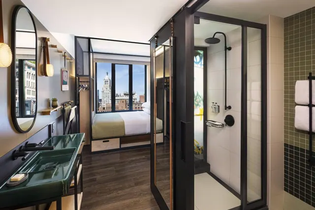 A hotel bathroom with a green vanity sink and a glass-enclosed walk-in shower, with a view through to a bedroom where a window shows a city skyline.