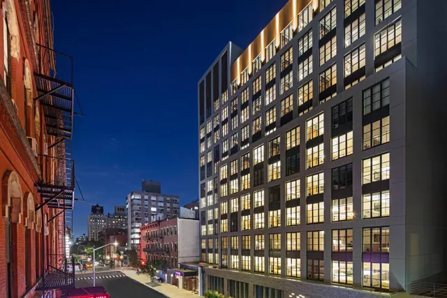 Dusk exterior view of a tall modern hotel facade with illuminated windows, beside an older red brick building on a city street.