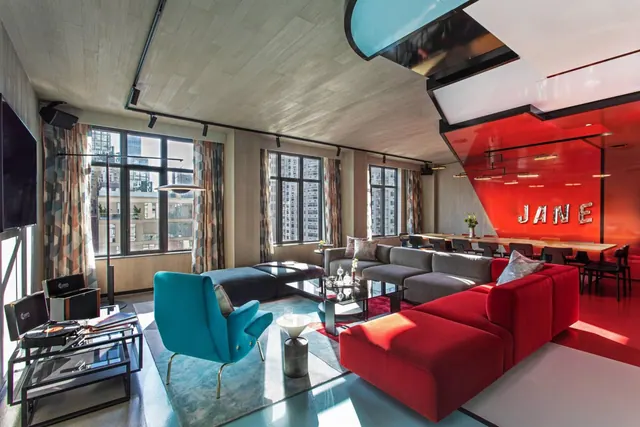 A living area with a red sectional sofa, a teal armchair, a television on the wall, city buildings visible through tall windows, and a red wall with the word JANE in illuminated letters.