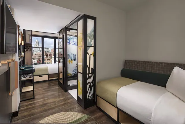 A hotel bedroom with a daybed in the foreground, a bed by the windows, a desk, a wall-mounted television, and a view of city buildings outside.