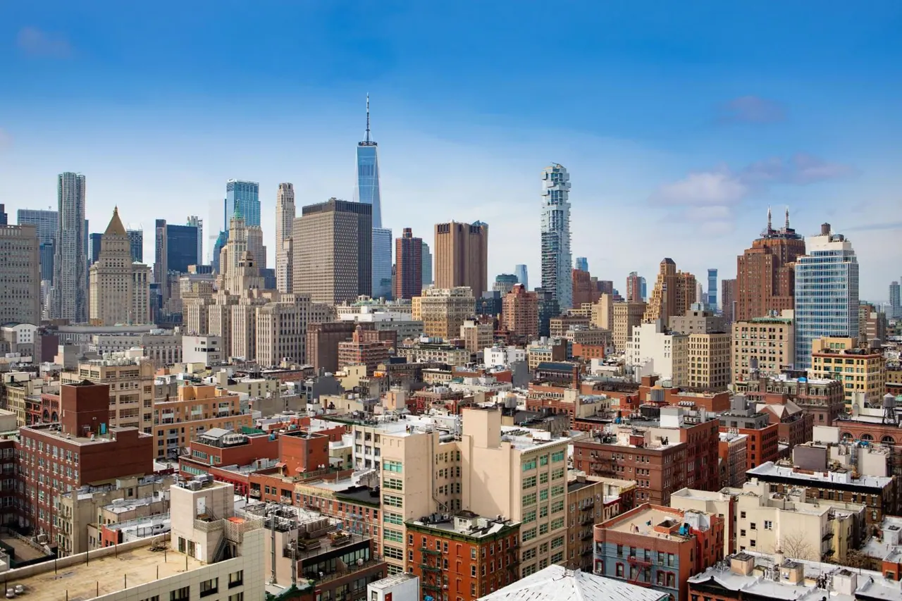 Elevated view over Lower Manhattan rooftops and skyscrapers, including One World Trade Center, under a blue sky.