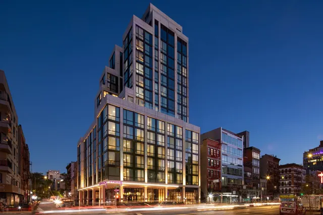 Exterior of a tall glass-and-stone hotel tower at dusk, with lit lower floors and a pink sign above the entrance, on a city street with traffic light trails.