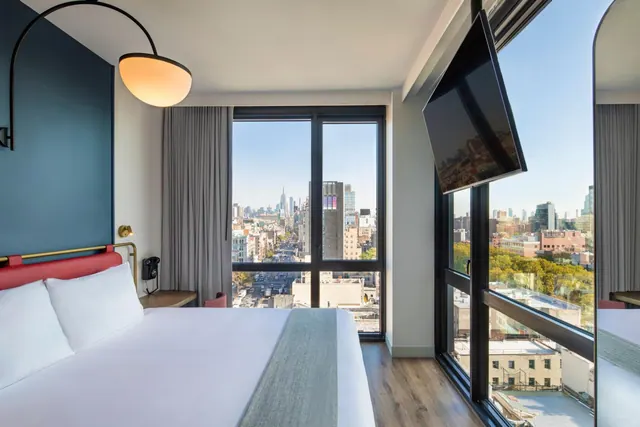 A king bed sits beside floor-to-ceiling windows, with a city skyline and the Empire State Building visible in the distance.