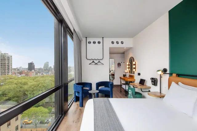 A hotel bedroom with a made bed, two blue armchairs, a desk with a laptop and floor-to-ceiling windows looking over a city street and buildings.
