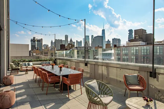 A rooftop terrace with a long dining table, orange chairs, lounge seating and string lights, with a city skyline including One World Trade Center in the background.