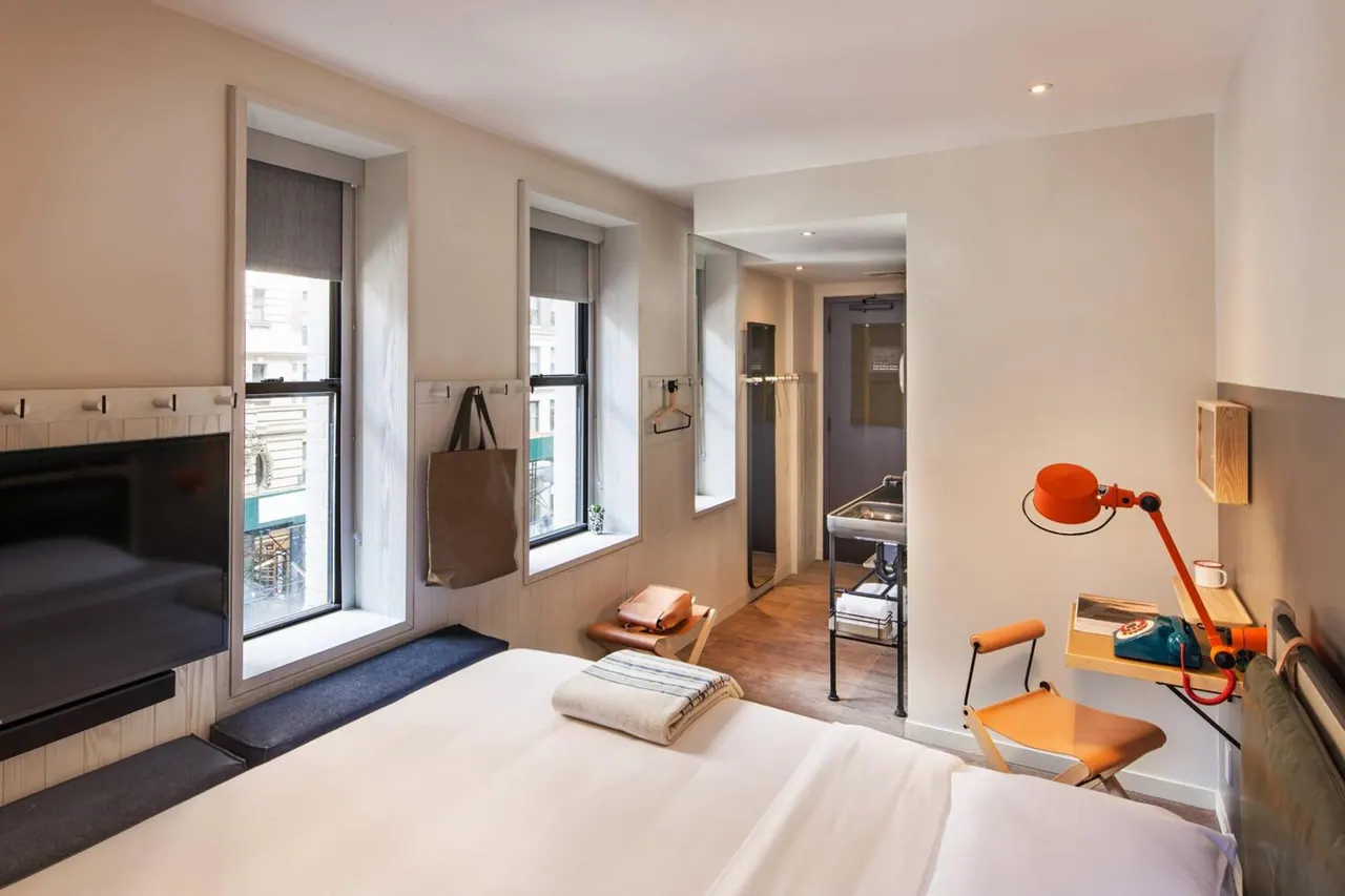 A bedroom with a made bed, a wall-mounted television, a wall desk with an orange lamp and chair, and two windows onto a city street, with a view through an open doorway to the bathroom.