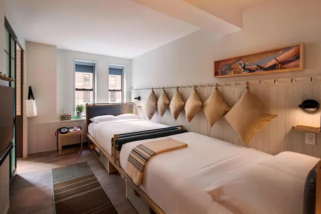 A bedroom with two twin beds against a wood-paneled wall hung with cushions, and windows looking onto a brick building.