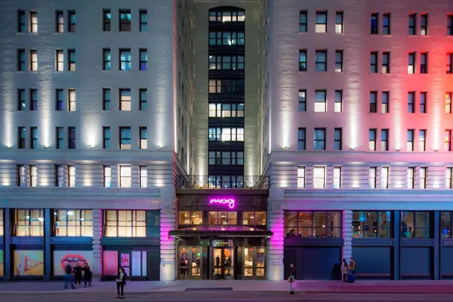 Night view of the hotel facade, lit with colored uplighting, with a pink neon sign above the entrance and a few people on the street.