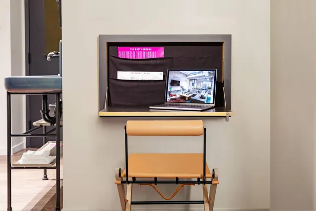 A wall-mounted fold-down desk holds an open laptop, with a leather chair in front and a pocket board of papers on the wall beside it.