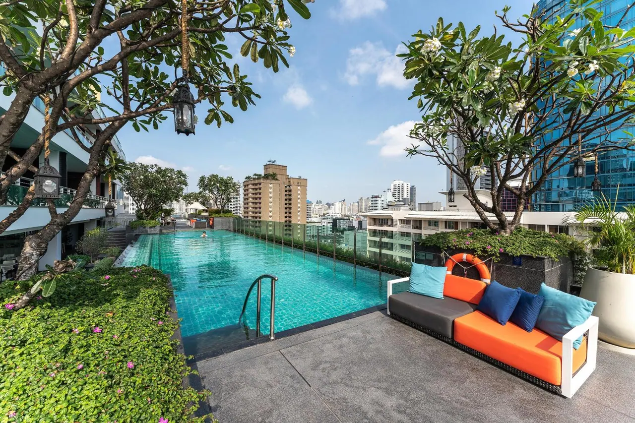 A rooftop swimming pool with an orange outdoor sofa and cushions in the foreground, frangipani trees overhead, and a city skyline with tall buildings in the background.