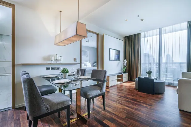 A staged living and dining area with a glass dining table, grey chairs, a low sideboard with a wall-mounted television, and floor-to-ceiling windows showing hazy high-rise buildings, with a bed visible through a doorway.