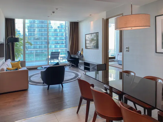 A living room with a beige sofa, round rug, black armchair, desk and dining table, with floor-to-ceiling windows looking onto a balcony and a glass high-rise opposite.